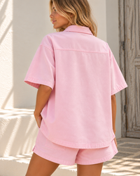 A woman wearing a light pink short-sleeve button-up shirt and matching shorts, shown from the back in a bright, sunlit outdoor setting.