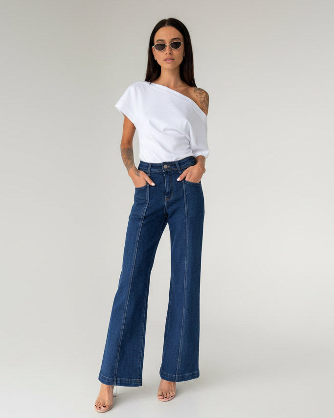A woman modeling a pair of dark blue wide-leg denim jeans with a white asymmetrical one-shoulder top, standing in a minimalist white studio setting.